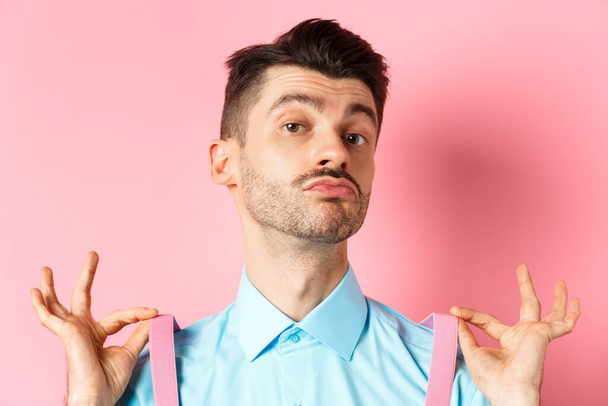 Close up of classy handsome guy adjusting suspenders, pucker lips and raise chin up, looking confident, standing in trendy outfit on pink background - Photo, Image