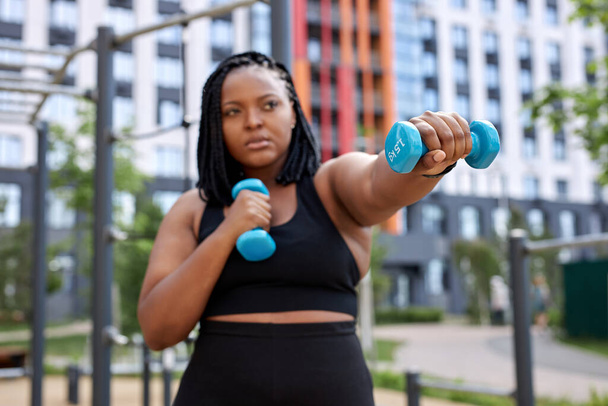 Chubby mixed race woman exercising with dumbbell in public park outdoors, concentrated - Foto, immagini