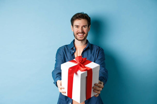 Valentines day. Handsome smiling man extending hands with gift box, wishing happy holiday. Guy making surprise present and looking cheerful, standing over blue background - Photo, Image