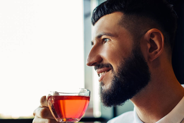 An attractive young bearded man, in a white T-shirt, sits in a cafe and drinks tea, a smiling guy holds a cup in his hand - 写真・画像