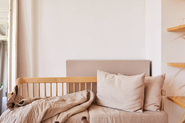 Фрагмент интерьера дома в натуральных пастельных тонах с деревянным диваном с мягкими бежевыми подушками и покрывалом, прикрепленным к белой стене - Фото, изображение