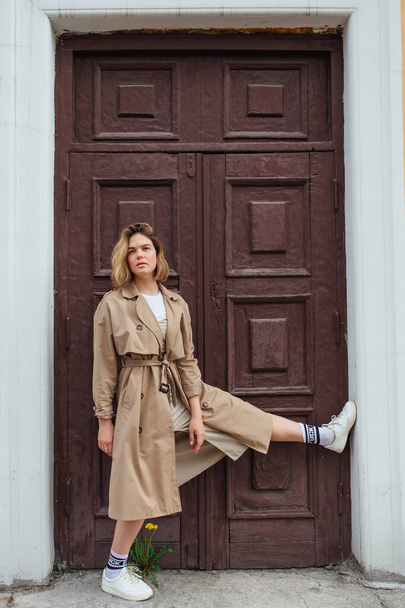 Young smiling millennial woman with wild hair dressed in an autumn coat posing near the door of an old building. - Foto, Imagen