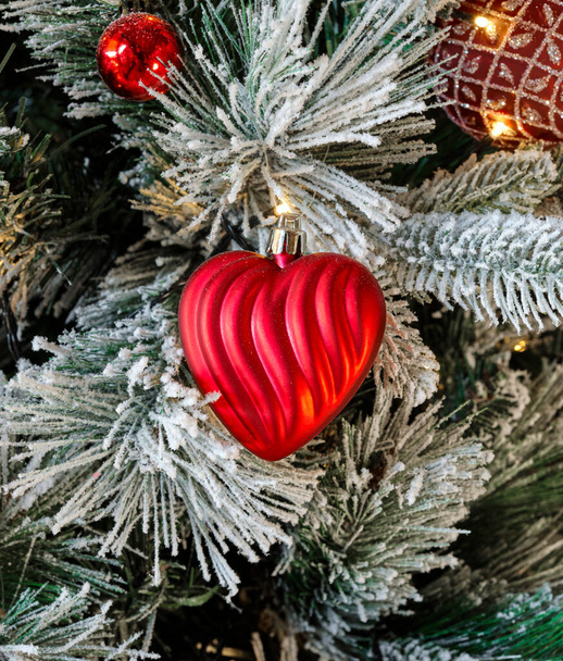 Dalları karla kaplı Noel ağacı ve kırmızı kalp şeklinde bir top, yılbaşı ışıklarıyla aydınlatılıyor. Gölgeler ve ışıklarla parıldıyor. Mutlu Noeller diliyor.. - Fotoğraf, Görsel