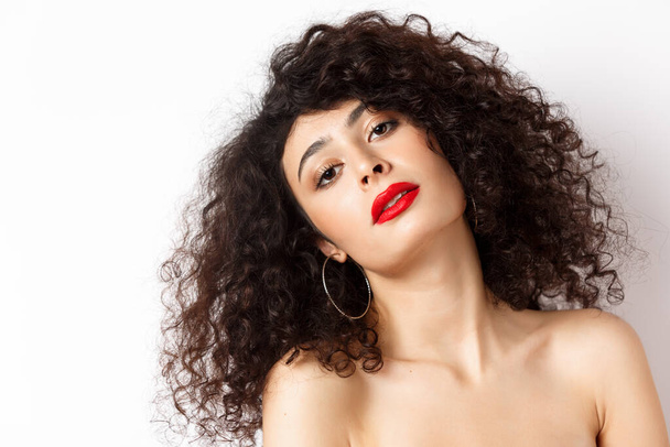 Close-up portrait of sensual and romantic woman with curly hair, red lips, looking seductive at camera, posing on white background - Photo, Image