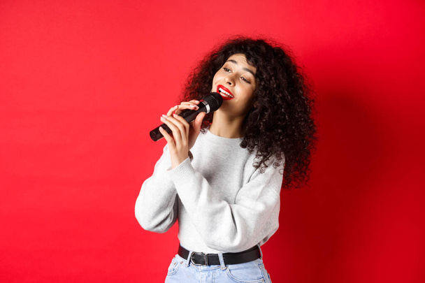 Leisure and hobbies concept. Stylish young woman singing karaoke, looking aside and holding microphone, performing song, standing on red background - Foto, afbeelding