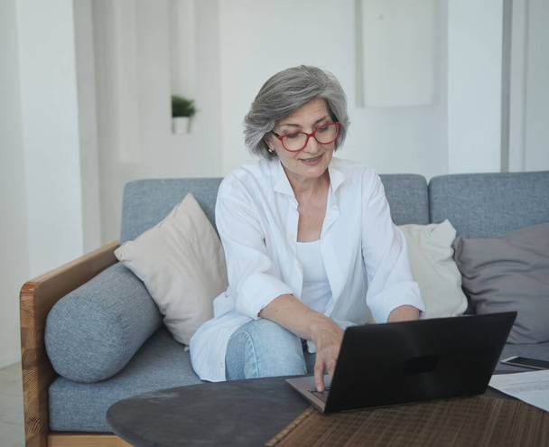 Προσεκτική ηλικιωμένη ανεξάρτητη γυναίκα που φοράει γυαλιά δακτυλογράφησης σε καναπέ για laptop. - Φωτογραφία, εικόνα