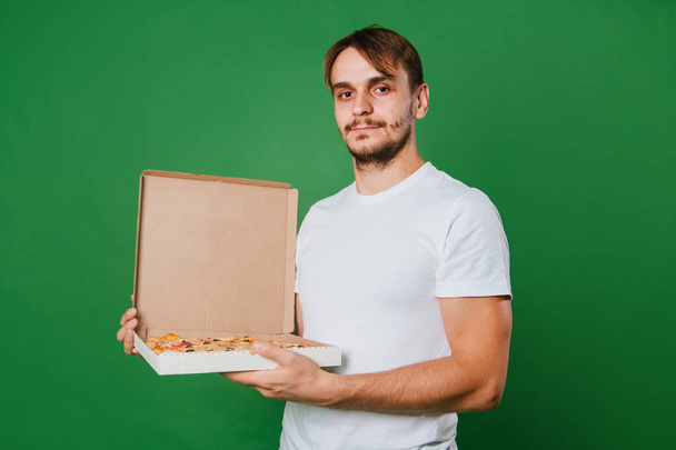 A young man in a white T-shirt holds a pizza box in his hands on a green background. Handsome guy Pizza delivery guy - Fotografie, Obrázek