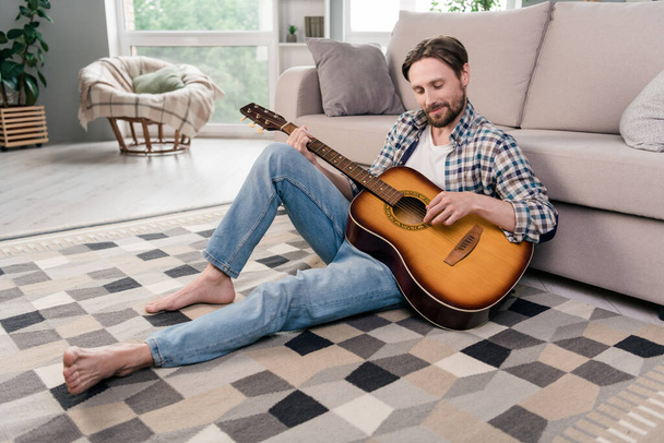 Photo of pretty handsome young man wear plaid shirt smiling sitting floor playing guitar indoors house room - Fotó, kép
