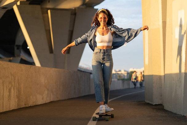 Молодая беззаботная женщина любит кататься на скейтборде на закате. Счастливая девушка в модной уличной одежде - Фото, изображение