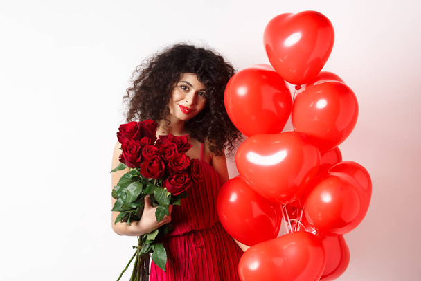 Wspaniałe kręcone włosy dziewczyna w sukni wieczorowej, posiadające datę, trzymając czerwone róże od chłopaka i pozowanie w pobliżu romantyczne serca balony, białe tło - Zdjęcie, obraz