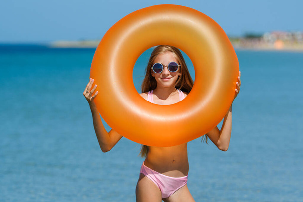 Portrait of a joyful girl on the beach with an orange rubber ring. The girl looks through the hole in the rubber ring. - Photo, Image