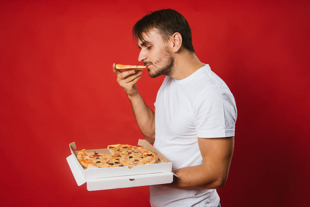 A young man in a white T-shirt holds a box in his hands and eats pizza. Handsome guy on a red background eats fast food.Mock-up. - Φωτογραφία, εικόνα