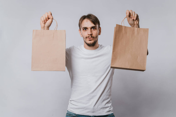 A young man in a white T-shirt holds clean paper bags in his hands. Handsome guy with purchases in his hands on a white background. Mock-up. - Foto, Bild
