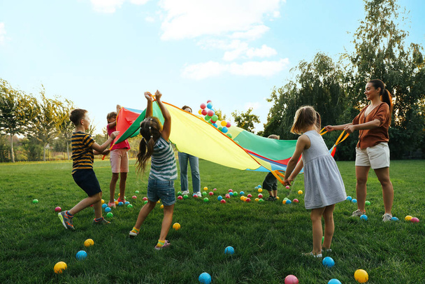 Group of children and teacher playing with rainbow playground parachute on green grass. Summer camp activity - Foto, Bild
