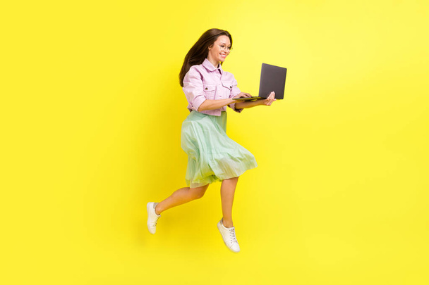 Full length body size photo portrait girl in skirt jumping high typing on laptop isolated bright yellow color background - Foto, imagen