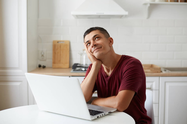 Handsome male wearing burgundy casual style t shirt sitting in front of portable computer at table in kitchen, dreaming about vacation, imagines how will be spend weekend. - Φωτογραφία, εικόνα