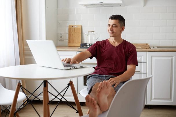 Positive man wearing burgundy casual t shirt sitting at table, putting feet on chair, using laptop for working or entertainment, having relaxed expression, browsing Internet or checking social networks. - Foto, Bild