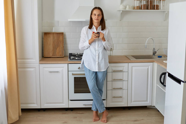 Full length portrait of woman drinking coffee in the morning at home, standing near kitchen set, enjoying hot beverage at home, wearing white shirt and jeans. - Φωτογραφία, εικόνα
