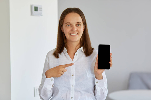 Mujer guapa con camisa blanca sosteniendo el teléfono inteligente en las manos y apuntando a su pantalla en blanco con el dedo índice, mirando a la cámara con una sonrisa encantadora. - Foto, imagen