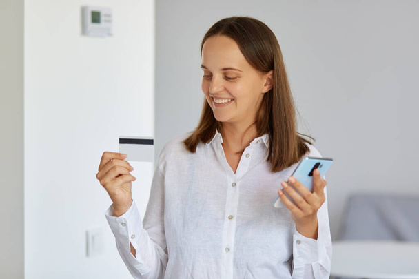 Indoor shot of positive satisfied female wearing white shirt standing at home, posing with smart phone in hand, showing credit card with big sum of money of on account. - Foto, afbeelding
