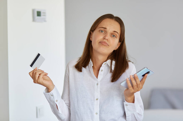 Portrait of puzzled female with dark hair standing with cell phone and credit card in hands, shrugging shoulders, does not know how spent all money from her bank card. - Foto, Imagem