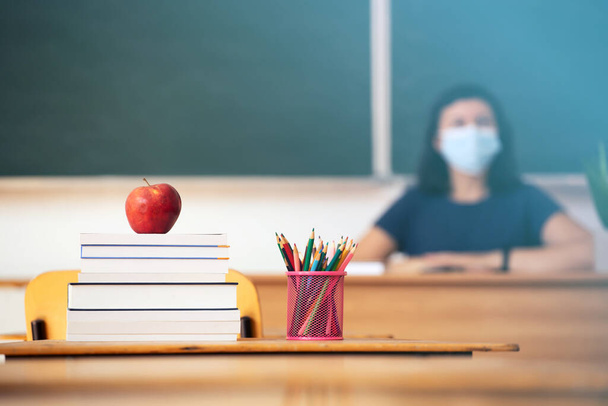 Back to School Concept. Apple, accessories and books in the classroom, teacher wearing mask in the background. - Φωτογραφία, εικόνα
