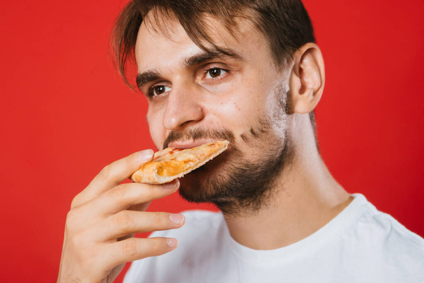 白いTシャツを着た若い男が手に箱を持ち、ピザを食べる。赤い背景にハンサムな男はファーストフードを食べます。. - 写真・画像