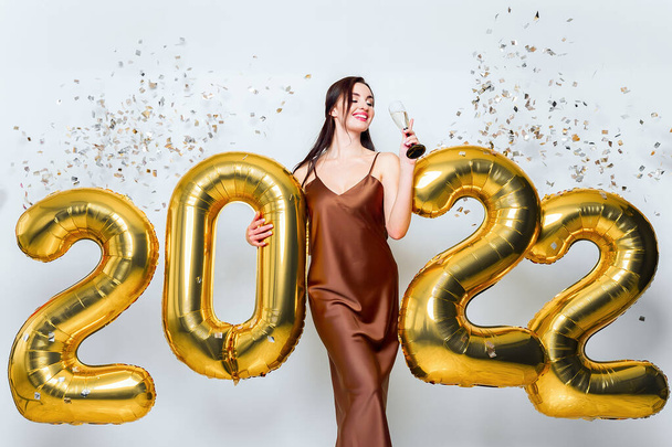 Happy young brunette woman with glass of champagne near golden 2022 balloons on white background with closed eyes. Confetti New Year celebration. Wearing fashionable glamor dress - 写真・画像