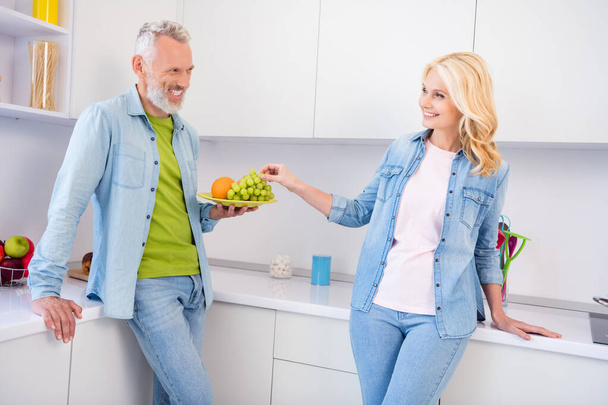 Photo of pretty shiny mature lovers dressed jeans shirt holding eating grape smiling indoors room home - Photo, Image