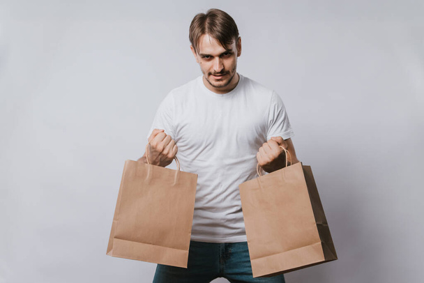 A young man in a white T-shirt holds clean paper bags in his hands. Handsome guy with purchases in his hands on a white background. Mock-up. - Фото, изображение