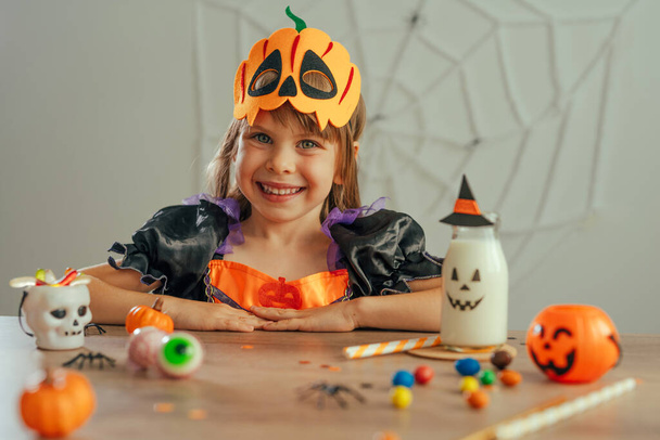 Porträt eines kleinen Mädchens im Halloween-Hexenkostüm und mit Kürbismaske am Tisch sitzend mit Süßigkeiten und Milch drinnen. Selektiver Fokus. - Foto, Bild