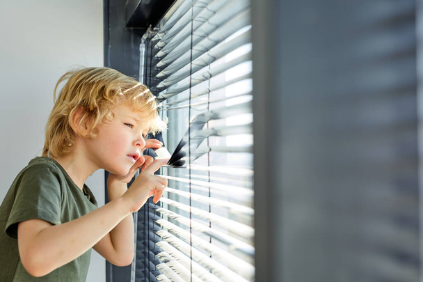 обиженный маленький мальчик смотрит в окно, опускает жалюзи, шпионов, мальчик остается дома - Фото, изображение