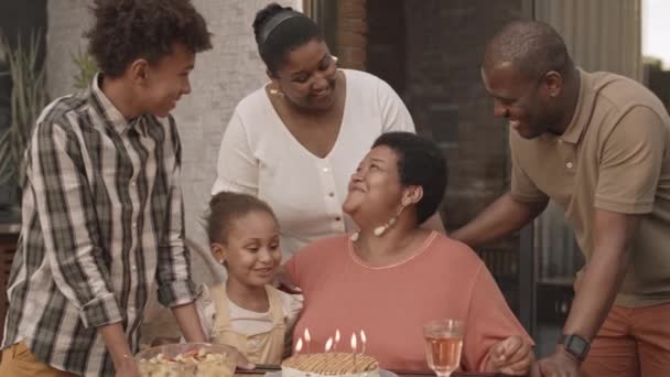成長したアフリカの女性の周りに立っている大人と若い子供の中長期テーブルに座っている彼らは上のろうそくと誕生日ケーキのための家族に感謝 - 映像、動画