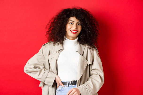 Modern woman with curly hair, wearing outdoor clothes, going for walk, standing against red background - Photo, Image