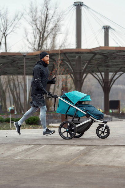 Motivated sportsman dad training and pushing baby carriage with a child in the park on a cold autumn day - Photo, image