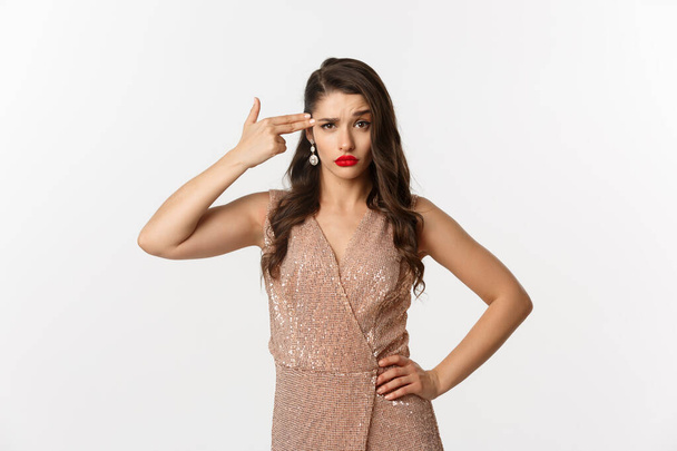 Celebration and party concept. Arrogant woman in elegant dress making finger gun at her head and looking bothered, standing annoyed over white background - Photo, Image