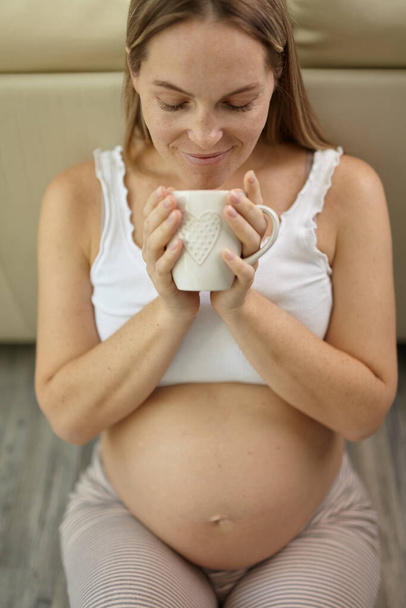 Portrait de femme enceinte caressant son ventre à la maison avec une tasse de café. Jeune femme enceinte blonde se sentant son bébé pousser, assis sur le sol, copier l'espace. - Photo, image