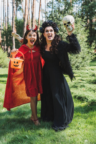 mom and daughter in halloween costumes holding bucket of sweets, skull and toy hand while screaming in forest - Photo, Image