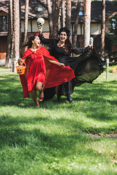 thrilled mom and daughter in halloween costumes running with skull, toy hand and bucket of candies - Фото, изображение