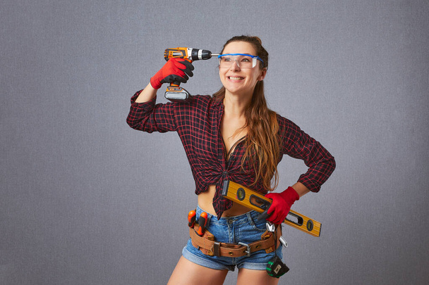 Young longhaired woman in casual clothes protective gloves hold electric drill or screwdriver in studio. Instruments accessories for renovation apartment. Repair home concept - Fotó, kép