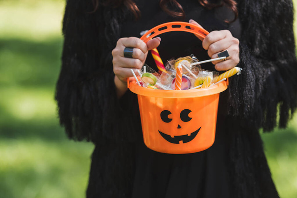 vista ritagliata della donna in abito nero che tiene secchio di Halloween con dolci - Foto, immagini
