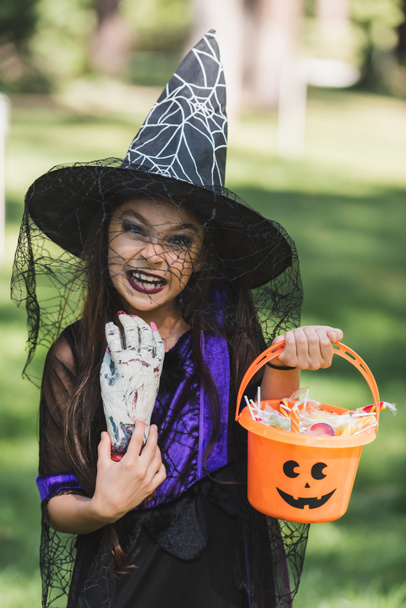 girl in witch hat making scary grimace while holding toy hand and bucket with candies - Foto, afbeelding