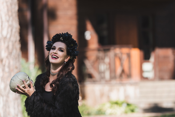 cheerful woman in vampire halloween costume holding spooky skull in backyard - Fotó, kép