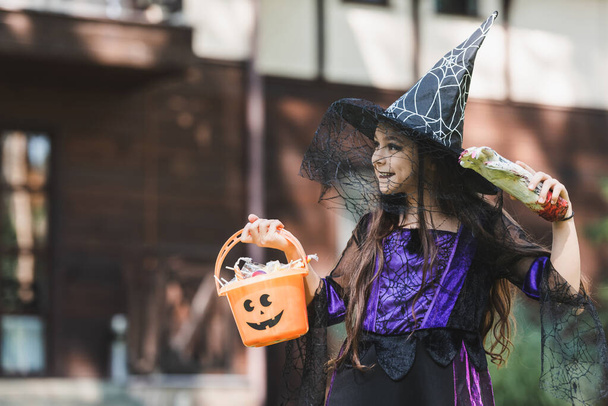 happy child in witch halloween costume, with toy hand and bucket with candies looking away outdoors - Photo, Image