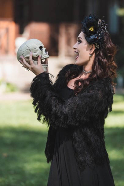excited woman in vampire halloween costume looking at spooky skull outdoors - Photo, image