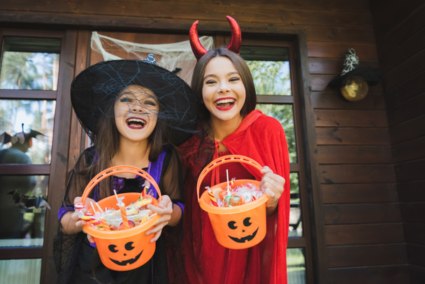 excited sisters in witch and devil halloween costumes holding buckets with sweets - Photo, Image