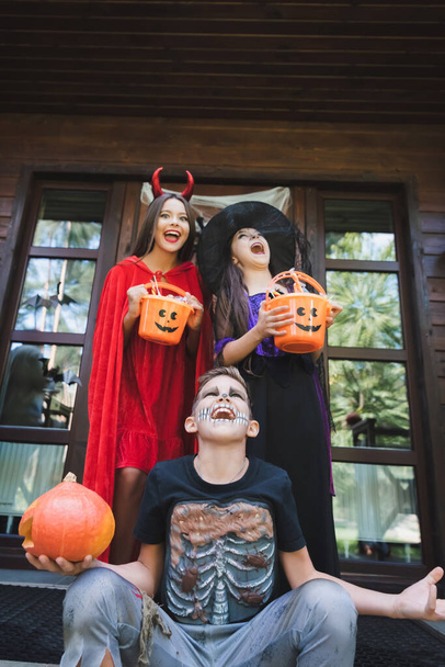 opgewonden kinderen in eng Halloween kostuums houden emmers met snoep en schreeuwen op de veranda - Foto, afbeelding