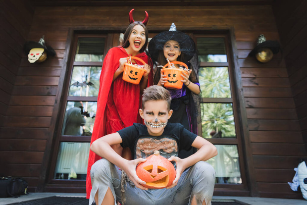 alegres hermanos en trajes de halloween, con calabaza tallada y cubos con caramelos en el porche de la casa - Foto, Imagen