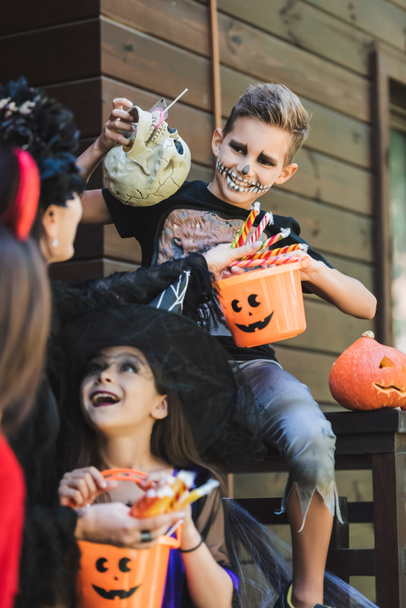 sonriente chico en esqueleto halloween traje celebración cráneo y cubo de dulces cerca borrosa mamá y hermanas - Foto, imagen