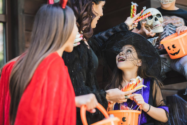 woman in halloween costume holding candies near amazed kids with buckets - Foto, imagen